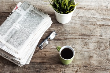 wooden tabletop with newspaper stack, glasses, a coffee mug, and a plant