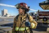 A woman in firefighting gear with a firetruck in the background looks in the direction of a structure that is being sprayed with water during a training exercise. 